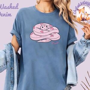 May include: A blue t-shirt with a pink snake illustration. The snake is coiled and decorated with heart shapes. The word "Love" is written in pink script below the snake. The shirt has the words "Washed Denim" in the upper left corner.