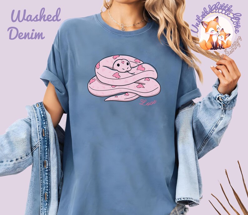 May include: A blue t-shirt with a pink snake illustration. The snake is coiled and decorated with heart shapes. The word "Love" is written in pink script below the snake. The shirt has the words "Washed Denim" in the upper left corner.