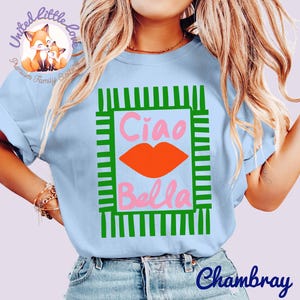 K&ouml;nnte beinhalten: Hellblaues T-Shirt mit einem gr&uuml;n-wei&szlig;en rechteckigen Design. Das Design zeigt die Worte "Ciao Bella" in Rosa, mit einer orangefarbenen Lippen-Grafik. Das Shirt hat kurze &Auml;rmel und ist aus einem weichen Material.