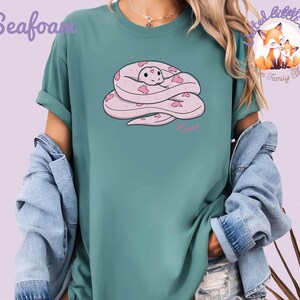 May include: Seafoam green t-shirt featuring a cartoon snake design. The snake is pink with heart-shaped accents and the word "Love" in pink script. The shirt has rolled-up sleeves and is worn with a denim jacket.