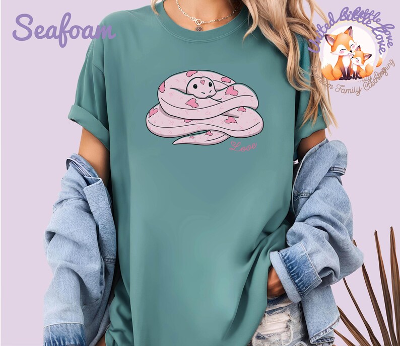 May include: Seafoam green t-shirt featuring a cartoon snake design. The snake is pink with heart-shaped accents and the word "Love" in pink script. The shirt has rolled-up sleeves and is worn with a denim jacket.