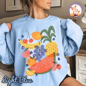 May include: Light blue sweatshirt featuring a colorful fruit graphic. The design includes a banana, cherries, lemon, pineapple, grapes, watermelon, orange slice, and strawberries. The words "Light Blue" are written in script at the bottom.