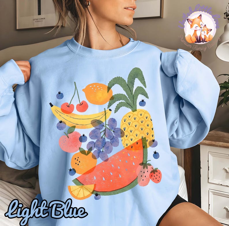 May include: Light blue sweatshirt featuring a colorful fruit graphic. The design includes a banana, cherries, lemon, pineapple, grapes, watermelon, orange slice, and strawberries. The words "Light Blue" are written in script at the bottom.