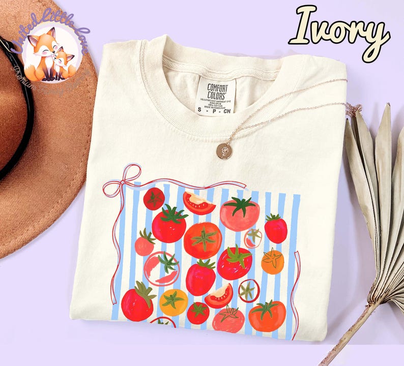 Peut inclure: T-shirt ivoire avec un motif de tomates. Le motif pr&eacute;sente des tomates rouges, des tranches de tomates et un fond &agrave; rayures bleues et blanches. Le t-shirt a un col rond et des manches courtes.