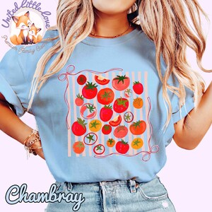 Peut inclure: T-shirt bleu clair avec un motif de tomates color&eacute;es. Le design pr&eacute;sente diverses tomates rouges, oranges et jaunes sur un fond ray&eacute;. Le mot "Chambray" est imprim&eacute; sur le t-shirt.