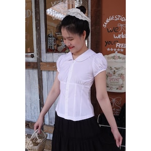 Puede incluir: Una blusa blanca de manga corta con cuello Peter Pan y corpiño entallado abotonado por delante. Se combina con una falda negra. También se ve un accesorio para el pelo blanco.