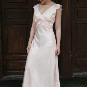 May include: A light pink, sleeveless dress with a V-neckline and a flowing skirt. The dress features a delicate floral pattern and ruffled trim along the bodice. The dress is made of a soft, silky material.