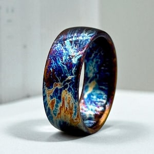 May include: A close-up of a colorful ring with a unique, textured surface. The ring features a blend of blues, oranges, and purples, creating a marbled effect. The ring is set against a white background.