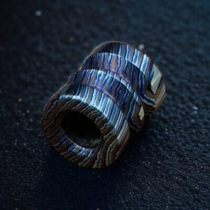 May include: A cylindrical bead with a layered pattern of blue, gray, and brown. The bead has a hole running through the center and is made of a material with a textured appearance. The design gives the bead a unique, artistic look.