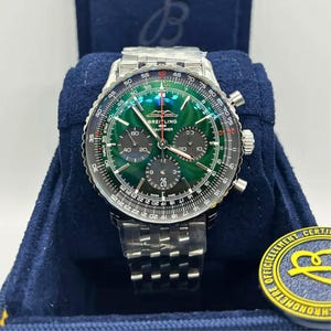 May include: A Breitling Navitimer watch with a green face and silver band. The watch has multiple sub-dials and a date display at the 6 o'clock position. The watch is presented in a blue box.