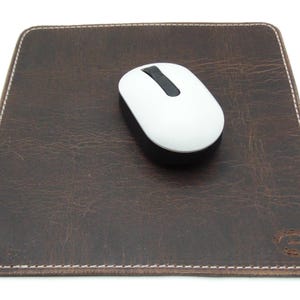 May include: A brown leather mousepad with white stitching and a white wireless mouse. The mousepad is rectangular and has a distressed texture. The mouse has a black scroll wheel.