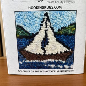 May include: A rug hooking kit featuring a sailboat on the water. The image shows a sailboat with white sails, brown hull, and blue water and sky. The kit is labeled "Schooner on the Bay" and measures approximately 6" x 6". The brand name "Deanne Fitzpatrick Studio" is visible.