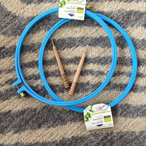 May include: Two bright blue plastic embroidery hoops with metal adjustment screws and white product labels reading embroidery hoop 25 cm 10 inches. Two wooden handled tools with thin metal tips are positioned inside the circular frames.