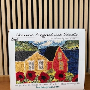 May include: A rug hooking kit featuring a colorful landscape scene with two houses and red poppies. The kit is labeled "Poppies on the Edge of Town" and measures 11" x 14". The box also includes the text "Deanne Fitzpatrick Studio" and "create beauty everyday".