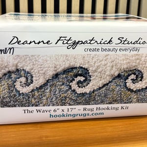 May include: A rug hooking kit titled "The Wave" by Deanne Fitzpatrick Studio. The kit features a wave design in shades of white, blue, and gray. The rug measures 6 inches by 17 inches. The text "create beauty everyday" is also visible.