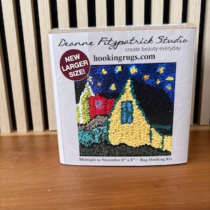 May include: A rug hooking kit featuring a colorful, stitched image of a house under a starry night sky. The kit is labeled "Midnight in November" and includes the text "hookingrugs.com". The box is marked "New Larger Size!"