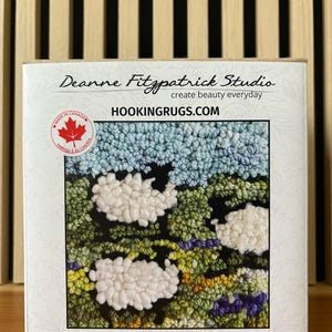 May include: A rug hooking kit titled "Sheep in Lavender" by Deanne Fitzpatrick Studio. The kit includes a design of white sheep in a field of green, blue, and purple, with the text "HOOKINGRUGS.COM". The rug measures approximately 6 inches by 6 inches.