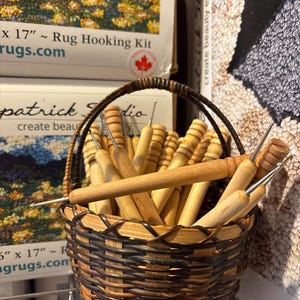 May include: A small, woven basket filled with wooden rug hooking tools. The basket is brown and black, with a handle. The tools have wooden handles and metal hooks. The background includes rug hooking kits.