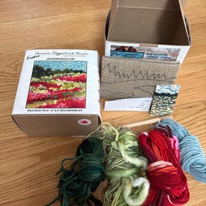 May include: A rug hooking kit with a box labeled "Wild Rose Field - 6" x 6" Rug Hooking Kit." The kit includes a box, yarn in various colors, a wooden tool, and a sample of the rug design. The box also has the text "Deanne Fitzpatrick Studio."