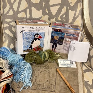 May include: A rug hooking kit with a puffin design, featuring a linen base, colorful yarn, and a wooden hook. The kit includes a box with the text "Deanne Fitzpatrick Studio" and "HOOKINGRUGS.COM".