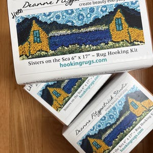 May include: Several rug hooking kits are stacked on a wooden surface. The kits are labeled "Deanne Fitzpatrick Studio" and feature a colorful landscape design with yellow houses and blue water. The text "Sisters on the Sea 6" x 17"" is also visible.