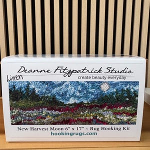 May include: A white box featuring a rug hooking kit with the text "Deanne Fitzpatrick Studio" and "New Harvest Moon 6" x 17" Rug Hooking Kit." The kit's image shows a landscape with a blue sky, trees, and a moon. The website "hookingrugs.com" is also visible.