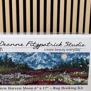 May include: A rug hooking kit featuring a landscape design with a moon. The kit is labeled "New Harvest Moon" and measures 6 inches by 17 inches. The box includes the text "Deanne Fitzpatrick Studio create beauty everyday".