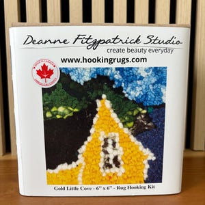 May include: A white box featuring a rug hooking kit with a colorful image of a yellow house with blue and green accents. The text on the box reads "Deanne Fitzpatrick Studio" and "Gold Little Cove - 6" x 6" - Rug Hooking Kit."