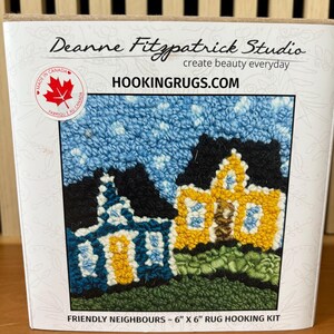 May include: A rug hooking kit featuring a colorful design of two houses with blue, yellow, and black details. The kit includes the text "Friendly Neighbours" and measures approximately 6 x 6 inches. The brand name "Deanne Fitzpatrick Studio" is visible.