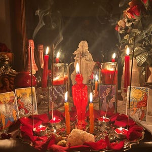 May include: A ritual altar display featuring a central red human-shaped candle, surrounded by multiple lit red and orange taper and tea light candles, incense sticks with rising smoke, scattered red rose petals, crystals, and tarot cards.