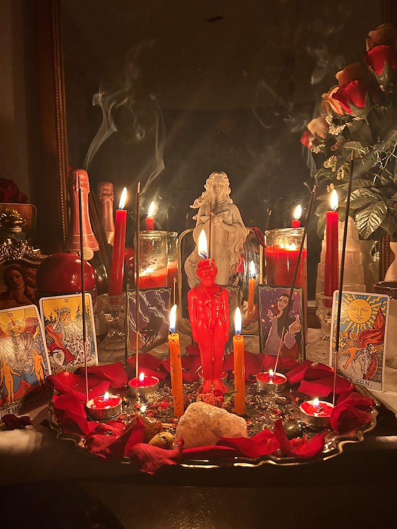 May include: A ritual altar display featuring a central red human-shaped candle, surrounded by multiple lit red and orange taper and tea light candles, incense sticks with rising smoke, scattered red rose petals, crystals, and tarot cards.
