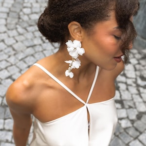 Pode incluir: Uma mulher usando brincos florais brancos e um vestido branco. Os brincos são compostos por flores de tecido branco e pequenos enfeites brilhantes. O vestido tem alças finas e um detalhe de fechadura.