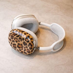 May include: White over-ear headphones with a leopard print ear cup cover. The ear cups are a light gray color, and the headband is white. The headphones are resting on a light-colored surface.