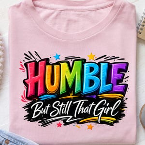 May include: A light pink t-shirt with the words "HUMBLE" in a rainbow gradient font, and the phrase "But Still That Girl" in a black script font. The design includes star accents and a black outline.