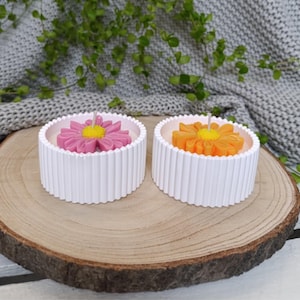 Könnte beinhalten: Zwei weiße, runde Kerzen mit geriffeltem Design. Jede Kerze hat eine blütenförmige Oberseite; eine ist rosa mit gelber Mitte, die andere orange mit gelber Mitte. Die Kerzen stehen auf einer Holzoberfläche.
