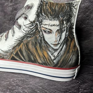 Op de afbeelding: Witte high-top sneaker met een handgeschilderd anime-stijl karakterportret. Het kunstwerk toont een gedetailleerd gezicht met donker haar en een bruine en beige jurk. De schoen heeft grijze veters en een rood-blauwe streep op de zool.