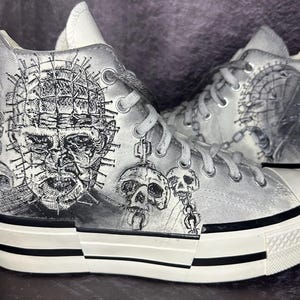 May include: Pair of high-top canvas sneakers with a black and white horror-themed design. The artwork features a detailed illustration of a character's face, skulls, and chains. The shoes have white laces, a white rubber sole, and a black stripe.