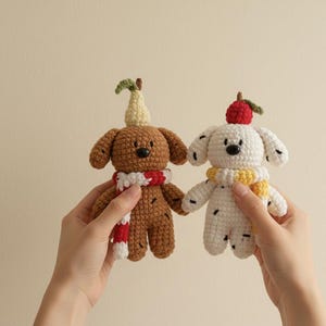 May include: Two handmade crochet dog figures. One is brown with a pear on its head and a red and white scarf. The other is white with black spots, a red apple on its head, and a yellow scarf. Both have black button eyes.