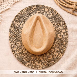 Puede incluir: Un sombrero de vaquero color beige con una corona marrón claro descansa sobre una superficie redonda estampada. La superficie presenta un diseño floral detallado en negro y beige. El texto "SVG - PNG - PDF | DIGITAL DOWNLOAD" está en la parte inferior.