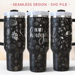 May include: Three black travel mugs with white lids and handles. Each mug is decorated with silver-colored illustrations and the words "IN MY SHOWGIRL ERA". The mugs have a variety of designs, including a strawberry, dice, and a cocktail.