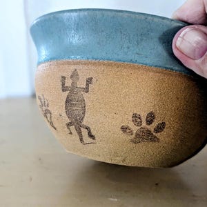 May include: A handmade ceramic bowl with a light blue rim and a tan body. The bowl features etched designs of a lizard and paw prints. The bowl is held by a person's hand.