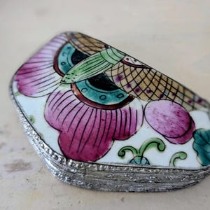 May include: A small, decorative trinket box with a hinged lid. The box features a floral design in shades of pink, green, and blue on a white background. The edges are trimmed with a silver-colored metal. The box is likely used for storing small jewelry items.