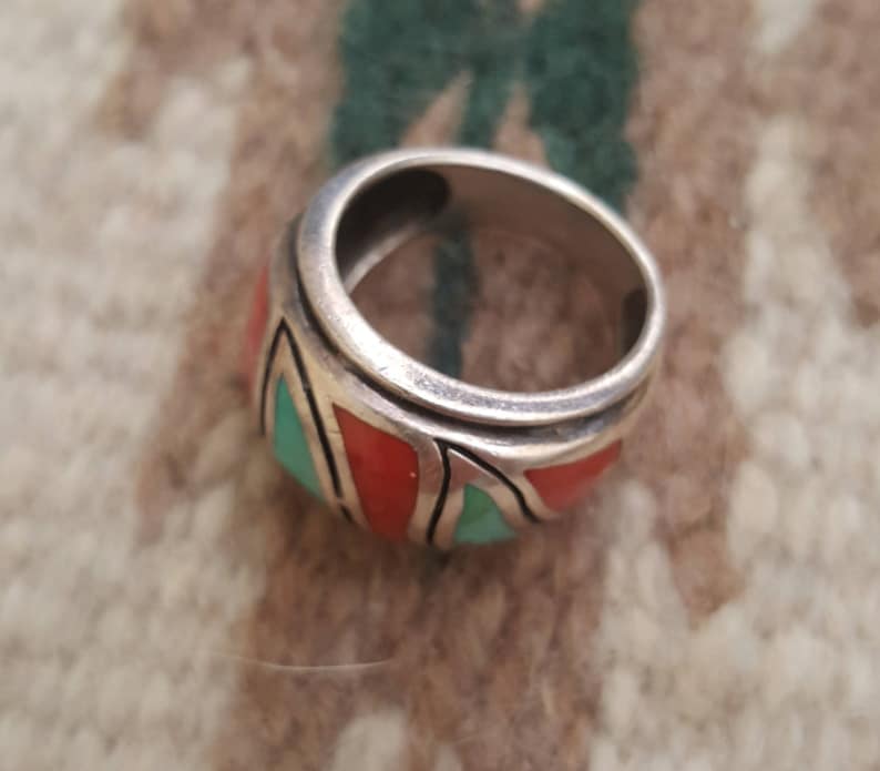 May include: A silver ring with a wide band featuring a geometric design inlaid with turquoise and coral stones.