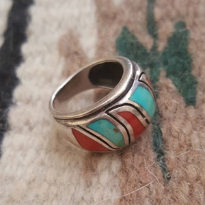 May include: A silver ring with turquoise and coral inlay. The ring has a wide band and a geometric design.
