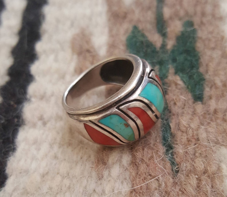 May include: A silver ring with turquoise and coral inlay. The ring has a wide band and a geometric design.