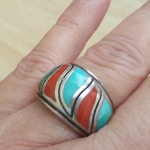 May include: A silver ring with turquoise and coral inlay. The ring has a wide band and a geometric design.