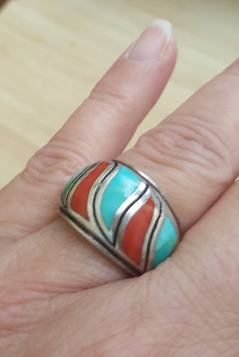May include: A silver ring with turquoise and coral inlay. The ring has a wide band and a geometric design.