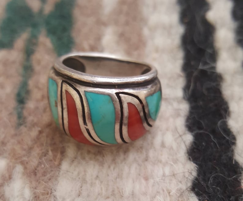 May include: A silver ring with turquoise and coral inlay. The ring is wide and has a rounded shape. The turquoise and coral are set in a silver band.