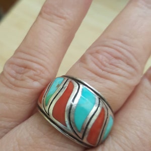 May include: A silver ring with turquoise and coral inlay. The ring has a wide band and a geometric design.