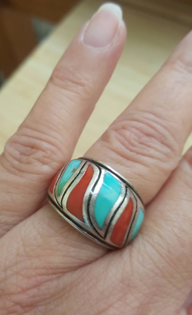 May include: A silver ring with turquoise and coral inlay. The ring has a wide band and a geometric design.